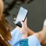 A young woman holding a smartphone while sitting outdoors in summer, capturing a moment of relaxation and technology use.